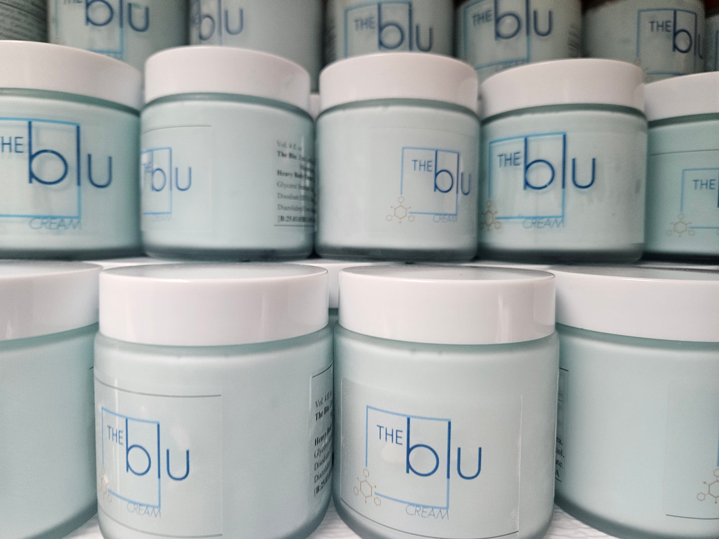 Several stacked light blue jars with white lids, labeled The Blu Cream - Zinc, Copper, and Magnesium in blue text, are closely arranged—this premium mineral cream is designed to soothe damaged skin.