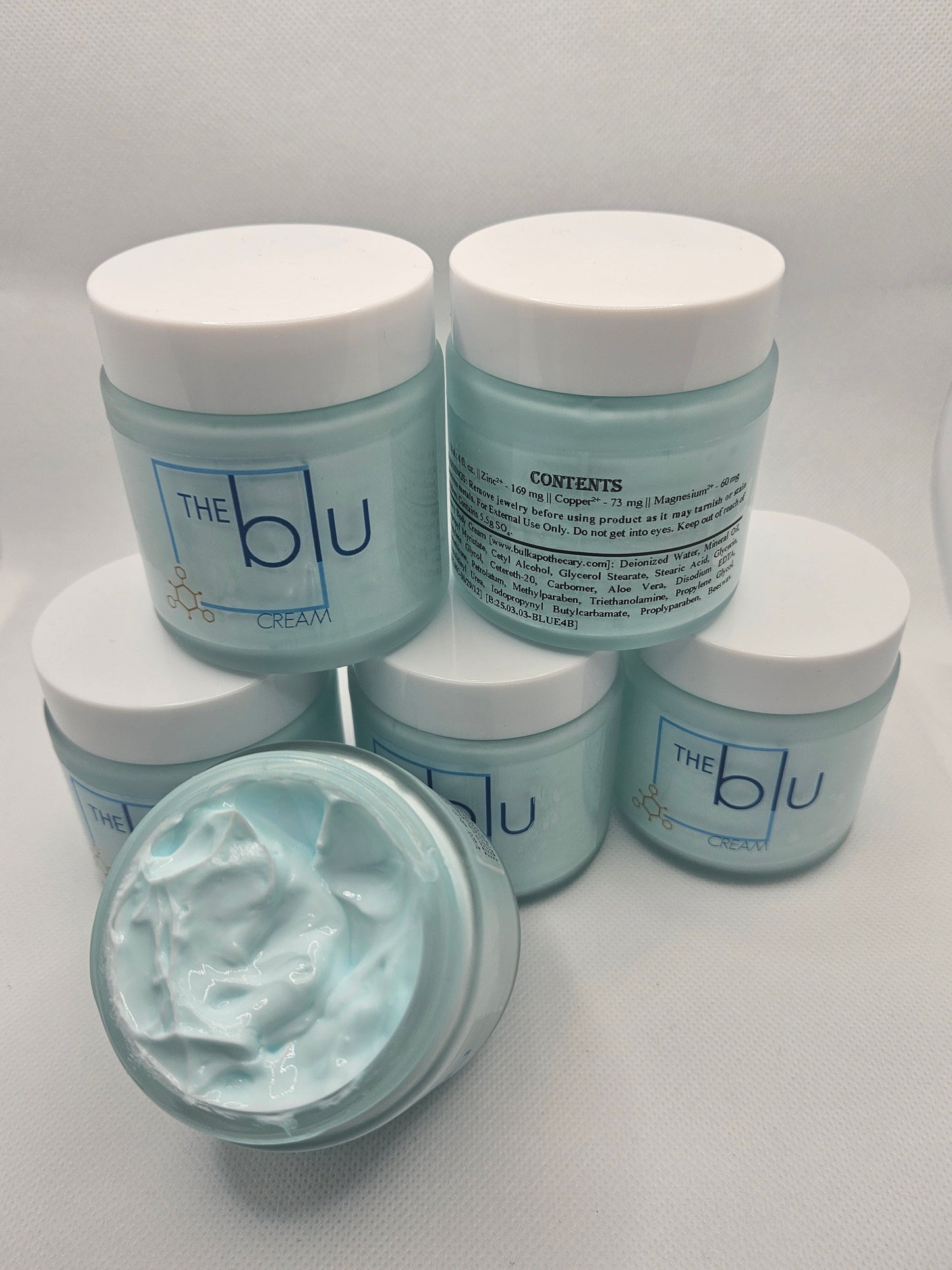 Five jars of The Blu Cream - Zinc, Copper, and Magnesium are shown on a white background. Four are closed; the open jar in front displays light blue cream inside, formulated to soothe damaged skin with zinc, copper, and magnesium.
