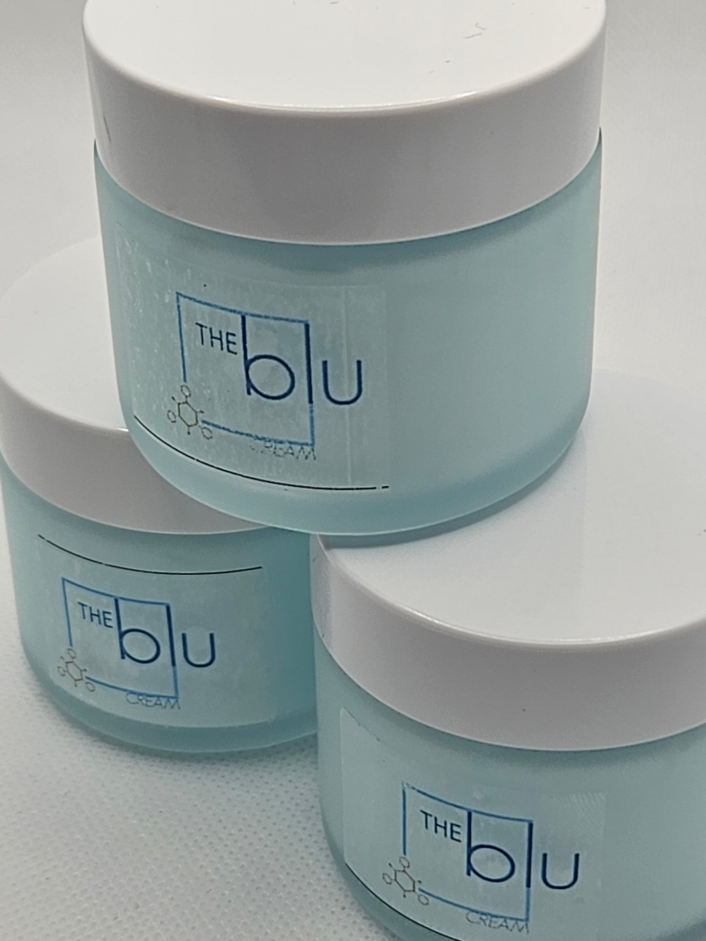 Three light blue jars with white lids are stacked together, each labeled The Blu Cream - Zinc, Copper, and Magnesium. With a simple blue and white design, this mineral cream soothes damaged skin. The background is plain white.