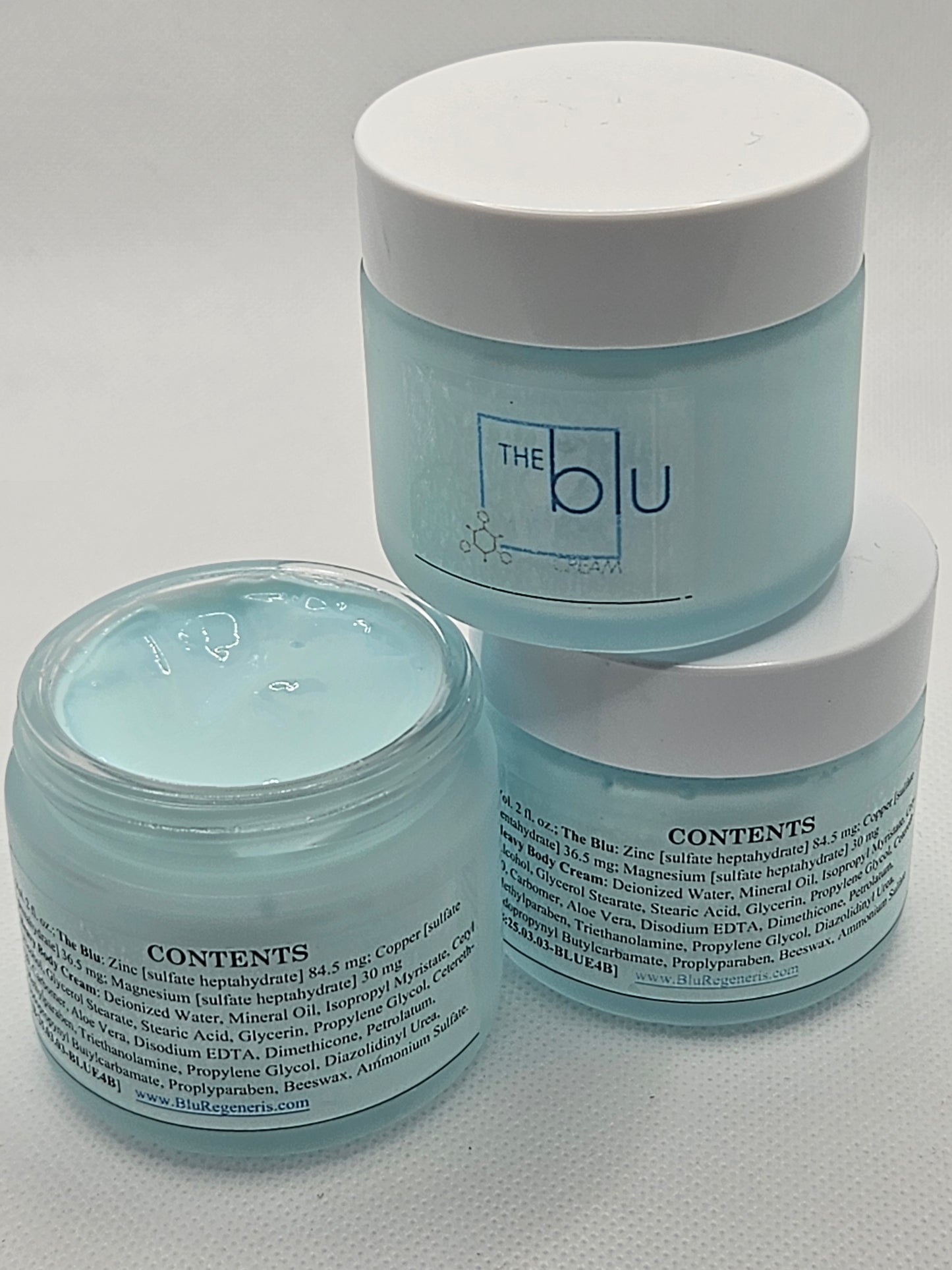 Three light blue jars labeled The Blu Cream - Zinc, Copper, and Magnesium are stacked on a white surface. An open jar shows pale blue cream, formulated to soothe damaged skin. Ingredient labels are visible.