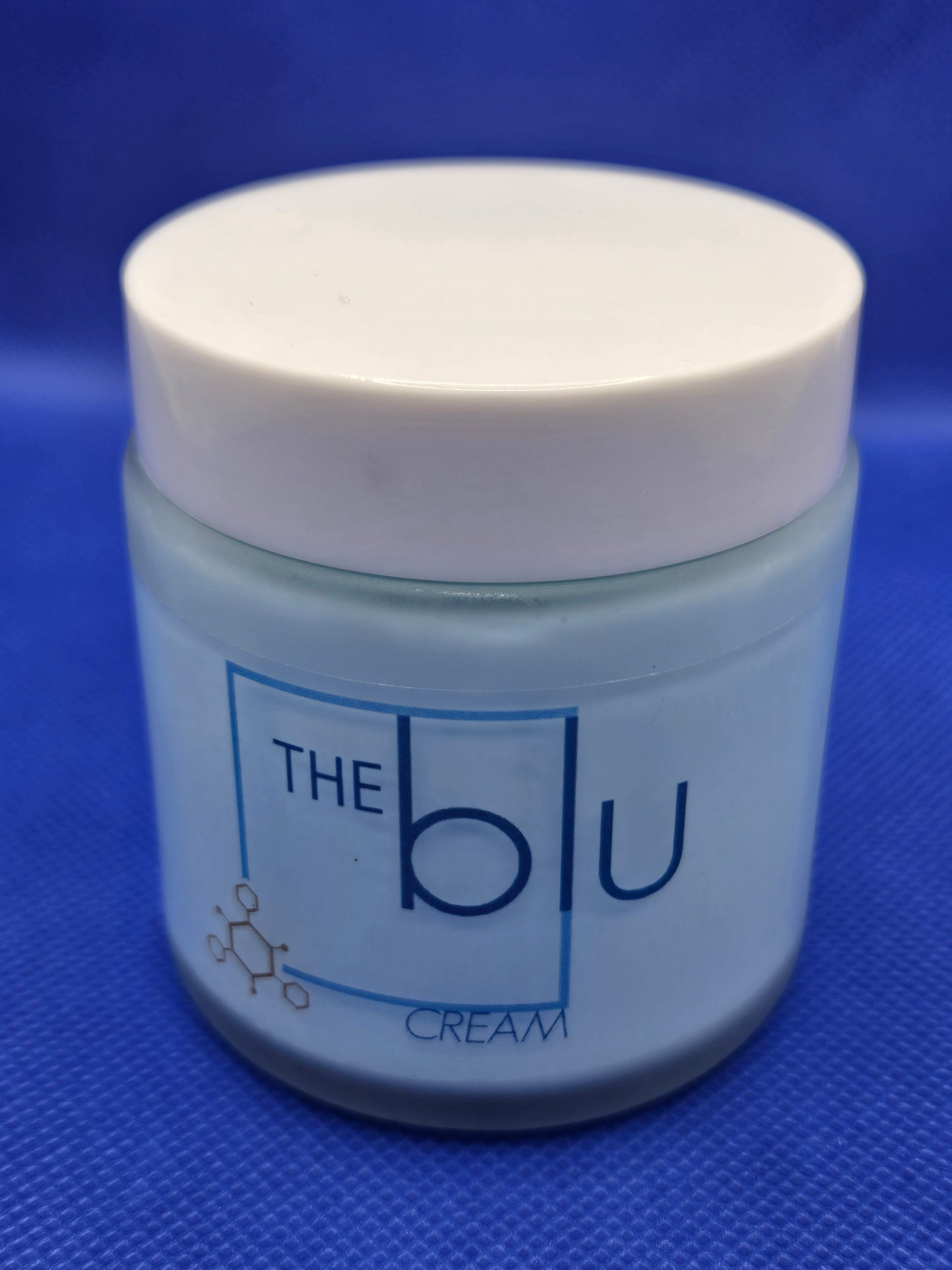 A light blue jar with a white lid labeled The Blu Cream - Zinc, Copper, and Magnesium, a mineral cream to soothe damaged skin, rests on a blue textured surface. A small molecular graphic is seen on the lower left of the label.