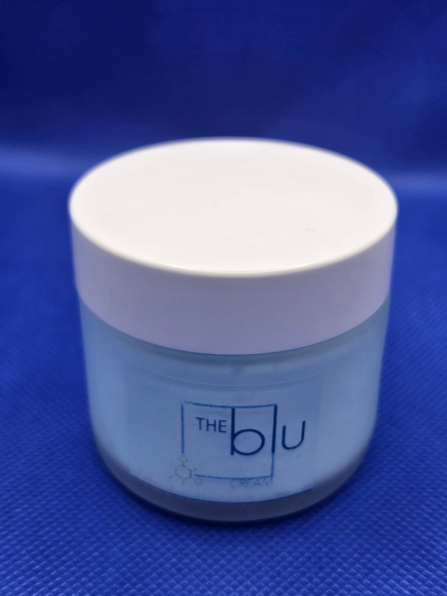 A small blue jar with a white lid labeled “The Blu Cream - Zinc, Copper, and Magnesium” rests on a textured blue surface; this mineral cream soothes damaged skin with zinc, copper, and magnesium.