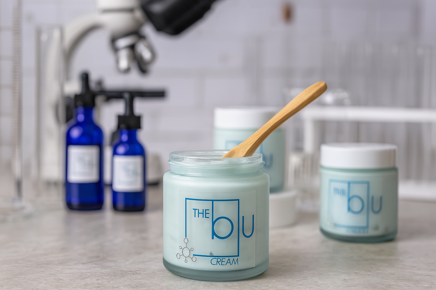 A glass jar labeled The Blu Cream - Zinc, Copper, and Magnesium sits on a counter with a wooden spatula. Blue dropper bottles and a microscope nearby evoke a lab setting for this mineral cream designed to soothe damaged skin.