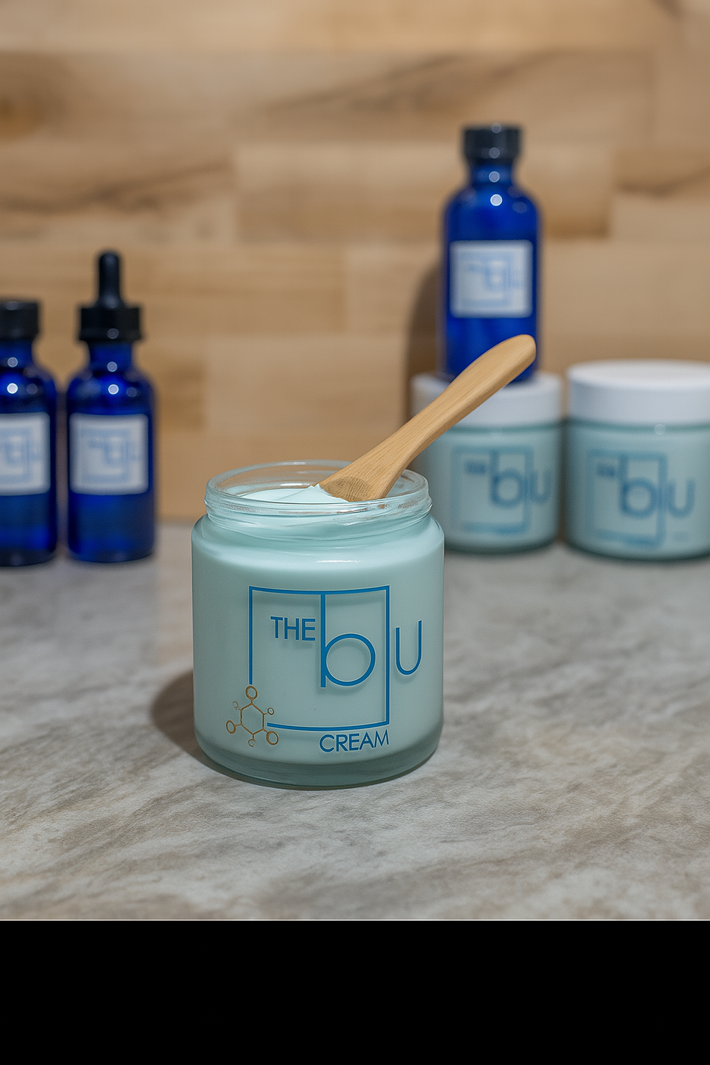 A jar of The Blu Cream - Zinc, Copper, and Magnesium rests on marble with a wooden spatula. Blue bottles and jars with matching labels stand in the background against a light wood wall.